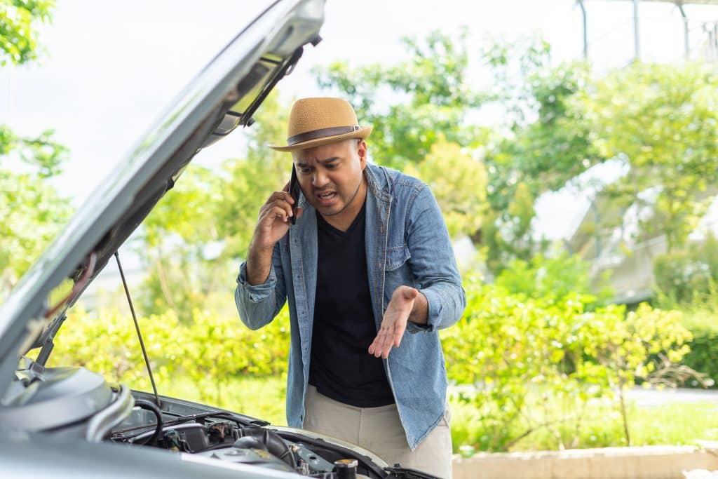 Frustrared young man looking down at car engine while on the phone.