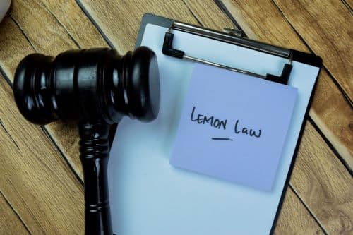A black gavel and clipboard with a blue sticky note that reads "Lemon Law" on a wooden table.