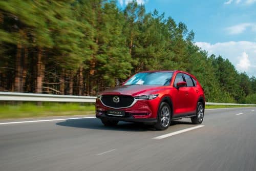 Red 2021 Mazda CX-5 driving on a road with trees in the background. 