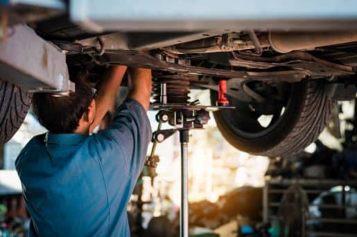 A blue-shirted man standing underneath a car and working on the transmission. 