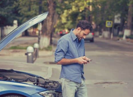 Upset man typing on his phone on the side of the road next to his broken-down car.