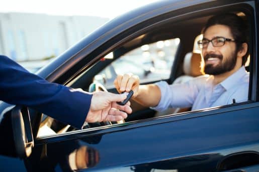 A man with glasses sitting in a used car, receiving keys from the salesman.